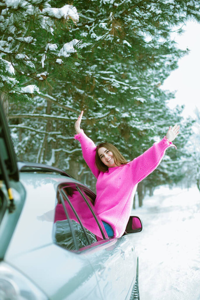 Young cute girl in a pink sweater laughs and looks out of the car window on a winter road in the snow  - 写真・画像
