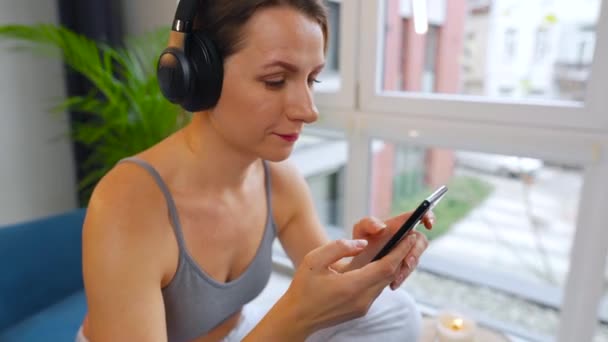 Woman in wireless headphones listening to music and using mobile apps or communicates on social networks on smartphone sitting on sofa at home - Filmati, video