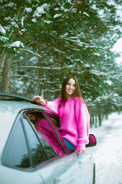 Young cute girl in a pink sweater laughs and looks out of the car window on a winter road in the snow  - Foto, imagen