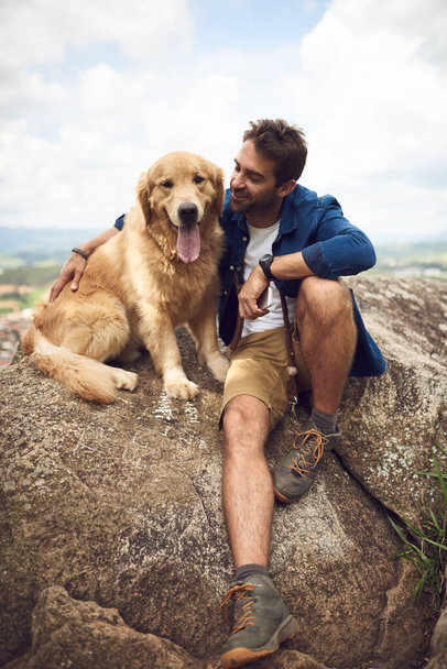 Well done boy, you made it. Full length shot of a handsome young man and his dog taking a break during a hike in the mountains. - Fotó, kép