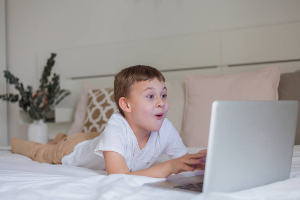 Adorable colegial utiliza Internet mientras está acostado en la cama en casa. Escuela en línea. Lindo niño de 7 años juega videojuegos en una computadora portátil. Acogedora casa. - Foto, Imagen