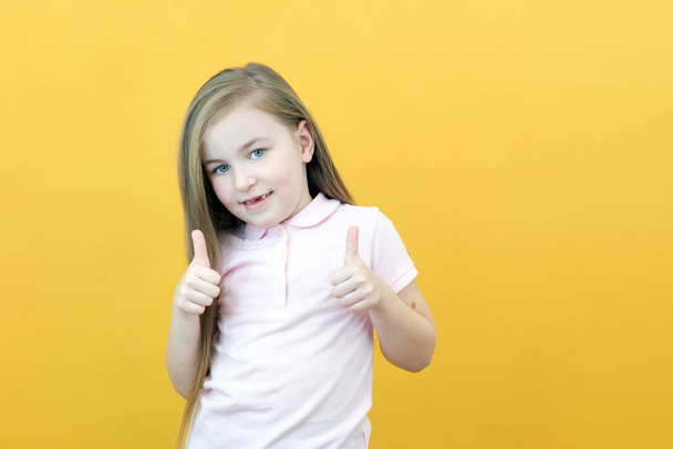Wow, look, advertise here! Portrait of an amazed cute girl pointing at an empty space in the background, a preschool girl shows a like on a yellow background - Fotoğraf, Görsel