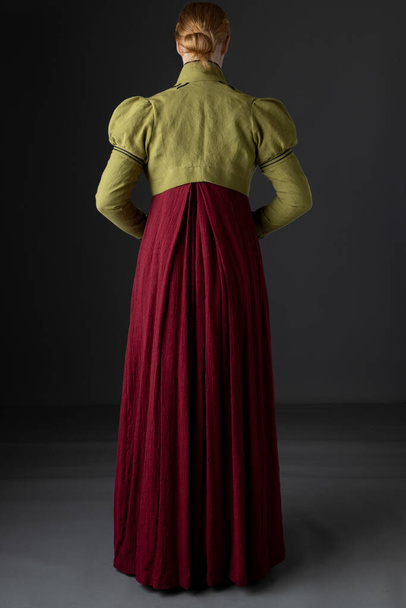 Una mujer regencia con un vestido de lino rojo con un spencer trenzado verde y guantes contra un fondo de estudio - Foto, imagen
