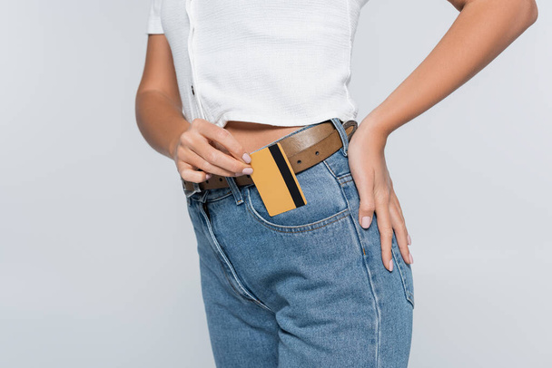 καλλιεργημένη άποψη της νεαρής γυναίκας βάζοντας πιστωτική κάρτα με cashback στην τσέπη σε τζιν που απομονώνονται σε γκρι - Φωτογραφία, εικόνα
