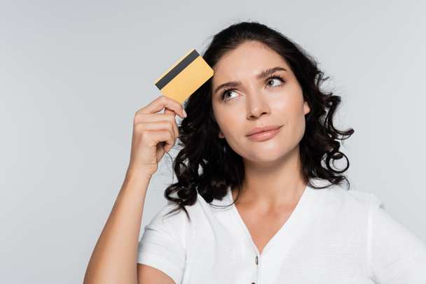 Nachdenkliche junge Frau hält Kreditkarte mit Bargeld isoliert auf grau - Foto, Bild