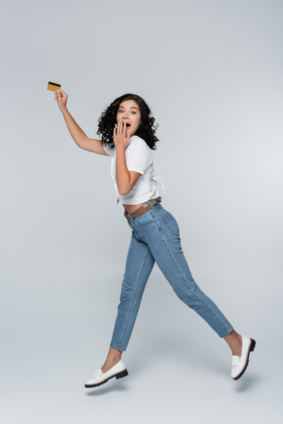 full length of amazed young woman in jeans holding credit card with cashback while levitating on grey - Фото, изображение