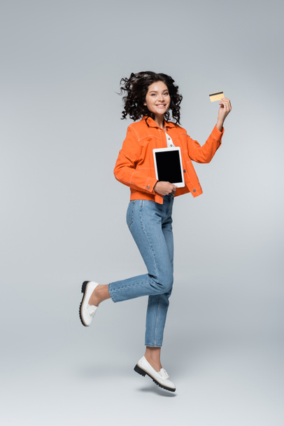 pełna długość szczęśliwej kobiety w pomarańczowej kurtce z kartą kredytową z kasą i tabletem cyfrowym podczas lewitacji na szarym - Zdjęcie, obraz