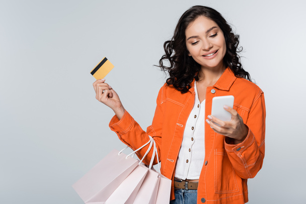 キャッシュバック付きのクレジットカードとグレーで孤立したショッピングバッグを持っている間にスマートフォンを使用して幸せな女性 - 写真・画像