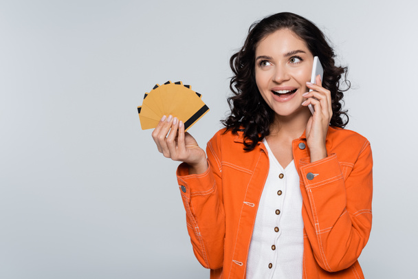 mujer joven feliz en chaqueta naranja con tarjetas de crédito con cashback y hablando en el teléfono inteligente aislado en gris - Foto, imagen