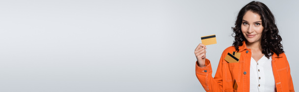 zadowolona młoda kobieta z kartami kredytowymi w kieszeni pomarańczowej kurtki odizolowana na szaro, baner - Zdjęcie, obraz
