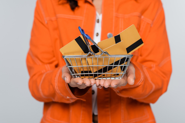 przycięty widok kobiety w pomarańczowej kurtce trzymającej koszyk zakupów z kartami kredytowymi odizolowanymi na szaro - Zdjęcie, obraz