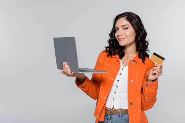 mujer joven y contenta con chaqueta naranja que sostiene el ordenador portátil y la tarjeta de crédito aislada en gris  - Foto, imagen