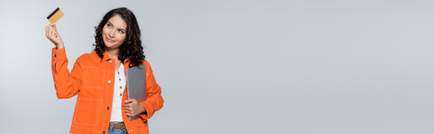 mujer complacida en chaqueta naranja sosteniendo el ordenador portátil y mirando la tarjeta de crédito aislada en gris, pancarta - Foto, Imagen