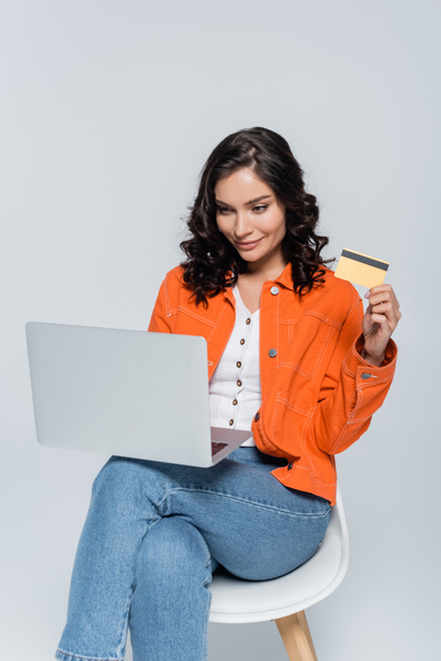 pleased young woman in orange jacket using laptop and holding credit card isolated on grey  - Zdjęcie, obraz