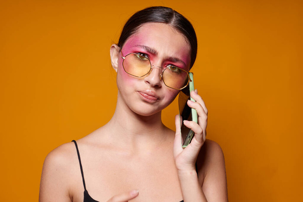 Sexy brunette woman with phone pink face makeup posing attractive look glasses yellow background unaltered - Фото, изображение