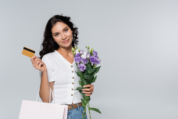 pleased woman holding bouquet of flowers, shopping bag and credit card isolated on grey  - Фото, зображення
