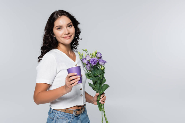 heureux jeune femme tenant bouquet de fleurs violettes et tasse en papier isolé sur gris  - Photo, image