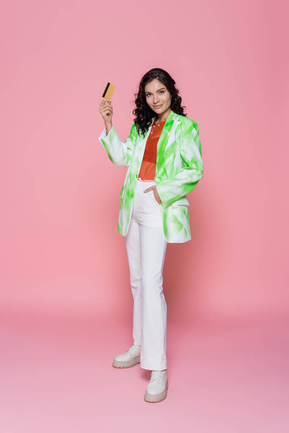 full length of pleased young woman in tie-dye blazer holding credit card and posing with hand in pocket on pink  - Zdjęcie, obraz