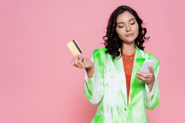 jonge vrouw in tie dye blazer houden credit card en smartphone geïsoleerd op roze  - Foto, afbeelding