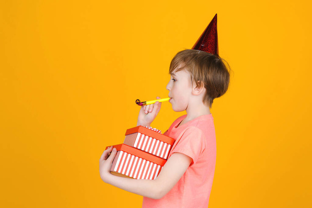 Tijd voor het feest. Vreugdevolle jongen met een feestelijke pet viert zijn verjaardag. Fluit blazen op een gele achtergrond. Gelukkige jongen met verjaardagscadeaus. Plezier, vreugde, feest en vakantie. Kopieerruimte. - Foto, afbeelding