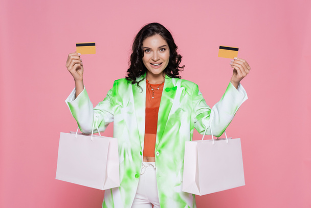 joyeuse jeune femme en blazer de teinture cravate tenant des cartes de crédit et des sacs à provisions isolés sur rose  - Photo, image