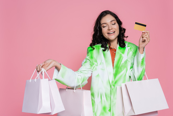 cheerful young woman in tie dye blazer holding credit card and shopping bags isolated on pink  - Zdjęcie, obraz