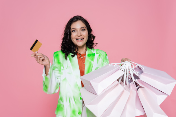 feliz joven mujer en corbata blazer tinte celebración de tarjeta de crédito y bolsas de compras aislados en rosa  - Foto, imagen