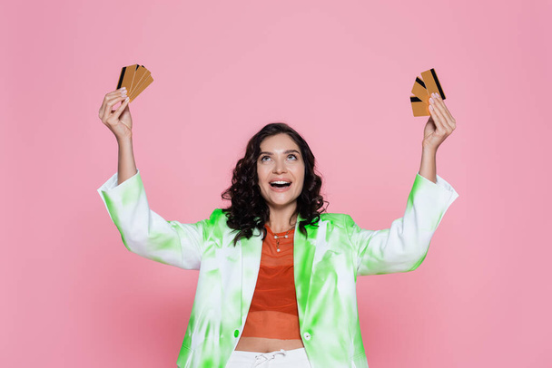 amazed young woman in green tie dye blazer looking at credit cards and smiling isolated on pink - Foto, afbeelding