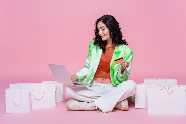 mujer complacida en blazer usando el ordenador portátil mientras sostiene la tarjeta de crédito cerca de bolsas de compras en rosa  - Foto, imagen