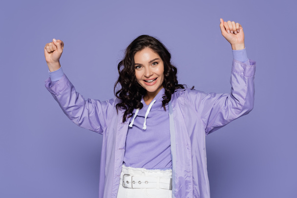 excited young woman in raincoat standing with raised hands isolated on purple - Foto, Imagem