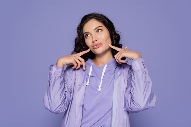 brunette young woman in raincoat pouting lips and pointing at cheeks isolated on purple - Photo, Image