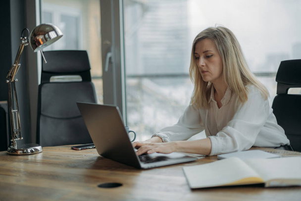 Уставшая женщина за своим столом в офисе. Молодая женщина с уставшим лицом, растрепанные волосы работает за компьютером на рабочем месте в течение дня - Фото, изображение