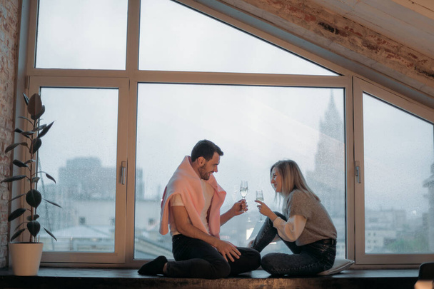 Mężczyzna i kobieta siedzą z kieliszkami szampana na parapecie przy oknie z widokiem na miasto. Kilku kochanków zorganizowało romantyczny wieczór, rozmawiając i pijąc szampana przy panoramicznym oknie w domu - Zdjęcie, obraz