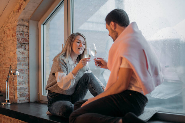 Bir adam ve bir kadın, pencerenin kenarındaki şehre bakan pencerenin kenarında şampanya kadehleriyle oturuyorlar. Bir çift aşık romantik bir akşam ayarladı, sohbet ediyorlar ve evdeki panoramik pencerede şampanya içiyorlar. - Fotoğraf, Görsel