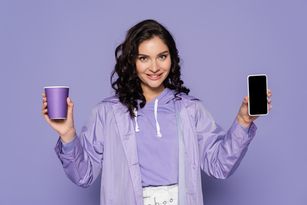 happy young woman in raincoat holding paper cup and smartphone with blank screen isolated on purple - Foto, Bild