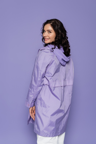 cheerful young woman in violet raincoat isolated on purple - Zdjęcie, obraz