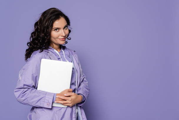 cheerful young woman in violet raincoat holding laptop isolated on purple - Foto, Imagen