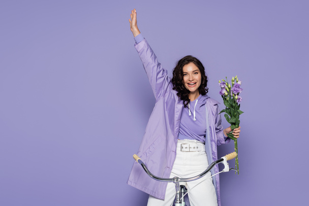 cheerful young woman in violet raincoat riding bicycle and holding flowers on purple - Photo, Image