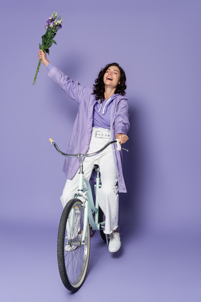 レインコートを着た幸せな若い女性が自転車に乗って紫色の花を持っています - 写真・画像