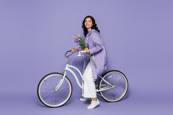 Mor yağmurluk giymiş, bisiklet süren ve elinde mor çiçekleri tutan neşeli genç bir kadın. - Fotoğraf, Görsel