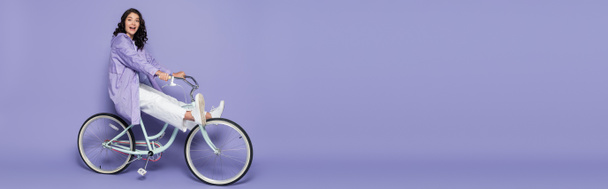 full length of amazed young woman in violet raincoat riding bicycle on purple, banner - Foto, Imagem