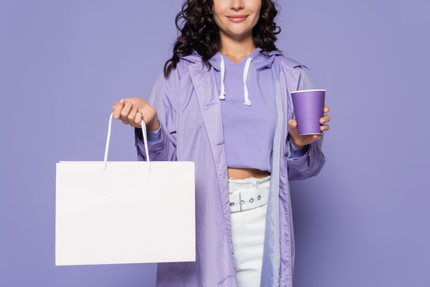 Yağmurluk giymiş, elinde kağıt bardaklar ve mor renkte izole edilmiş alışveriş torbası olan mutlu genç bir kadın manzarası. - Fotoğraf, Görsel