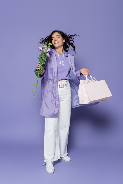 happy young woman with closed eyes holding shopping bags and flowers on purple - Foto, Imagen
