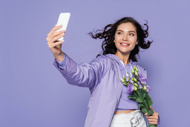 χαρούμενη νεαρή γυναίκα που κρατάει λουλούδια και βγάζει selfie στο smartphone που απομονώνεται στο μωβ - Φωτογραφία, εικόνα