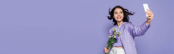 幸せな若い女性が花を持っていると紫、バナーに隔離されたスマートフォンで自撮り - 写真・画像