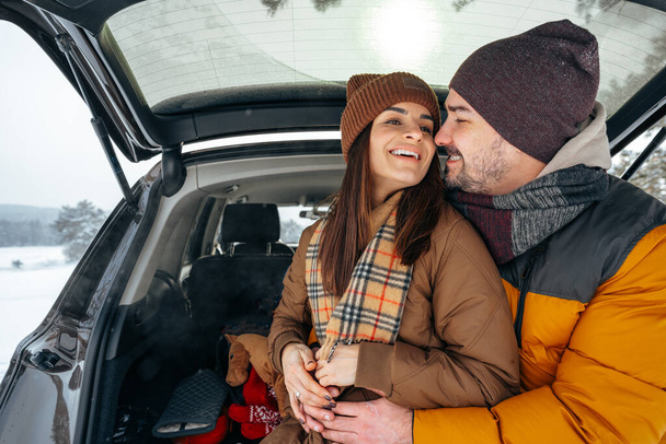 Lovely smiling couple sitting in car trunk in winter forest - Foto, afbeelding