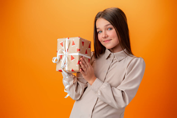 Cute teen girl holding a Christmas gift box against orange background. - Valokuva, kuva