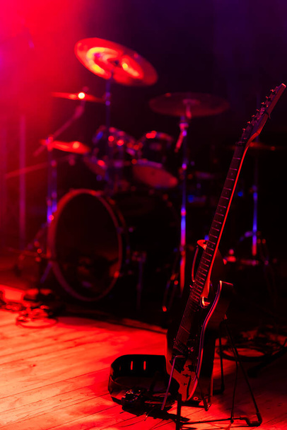 Μια μαύρη ηλεκτρική κιθάρα βασισμένη σε μια βάση τοποθετημένη στη σκηνή. Θέμα φωτίζεται από ισχυρό κόκκινο φως. - Φωτογραφία, εικόνα