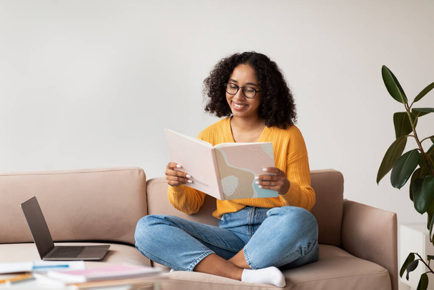 Радісна афроамериканська жінка, що сидить на дивані з ноутбуком і книжкою, виконує домашнє завдання, читає вдома, вільне місце - Фото, зображення