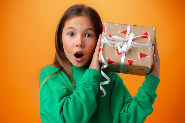 Cute teen girl holding a Christmas gift box against orange background. - Foto, afbeelding